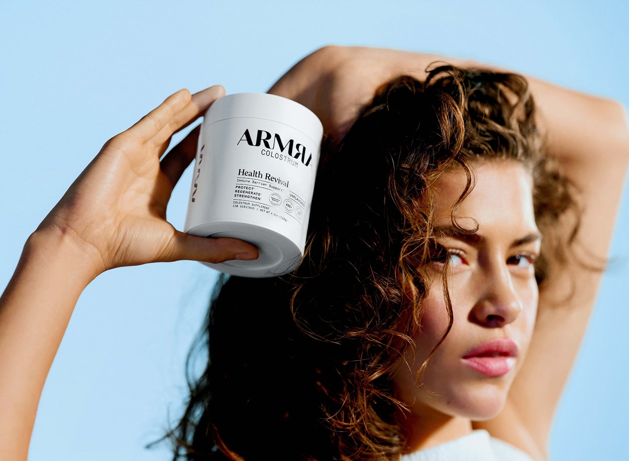 A woman with thick, curly brown hair holding a jar of ARMRA colostrum