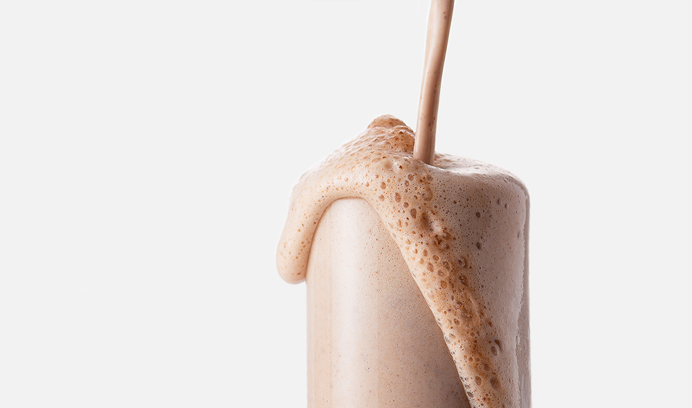 White background with a clear glass and ARMRA performance revival chocolate product being poured in