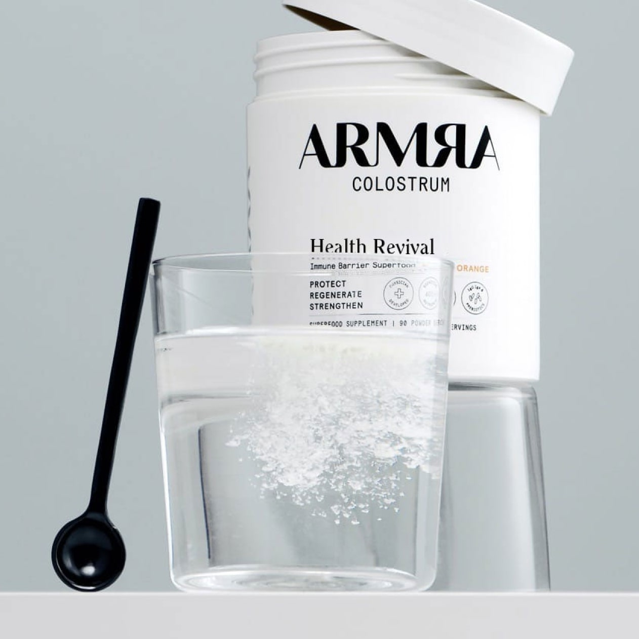 A glass of ARMRA colostrum next to a spoon and a jar of ARMRA colostrum