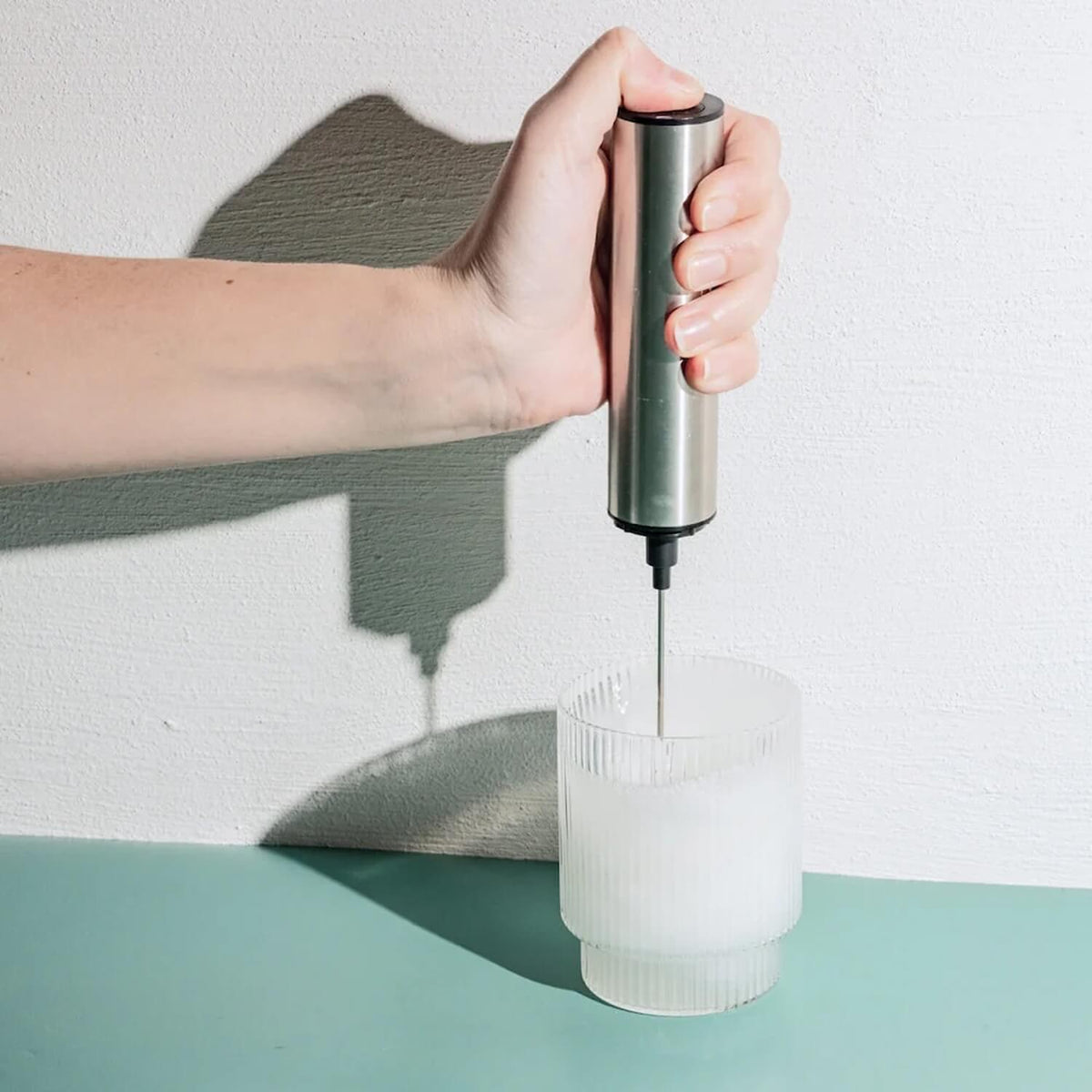 Hand holding stainless steel milk frother with whisk attachment positioned above white frother cup.
